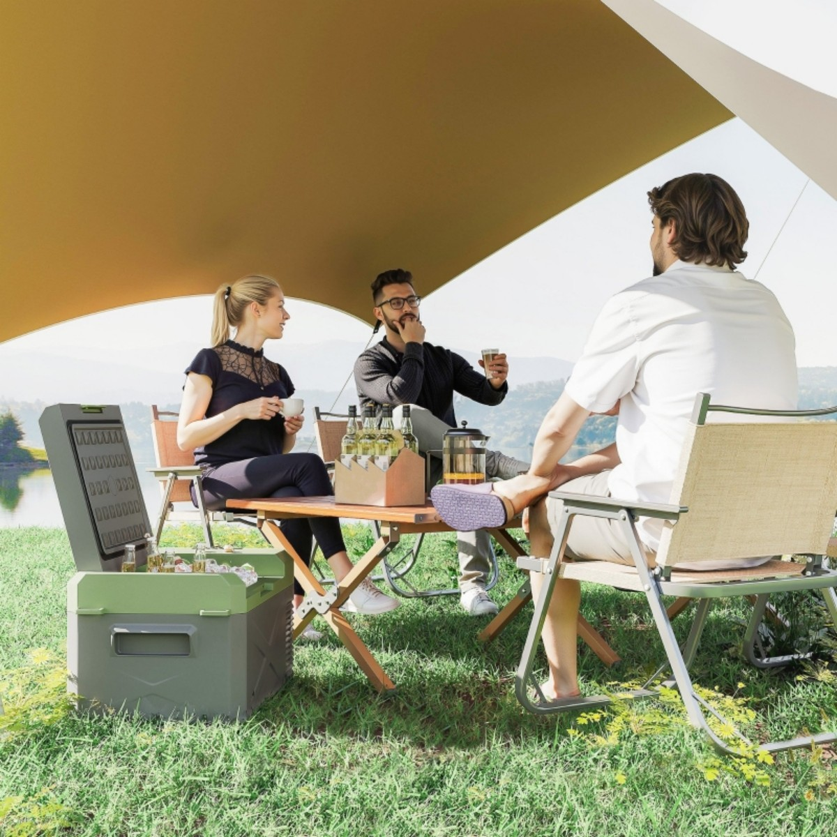 Drei Personen sitzen an einem Picknicktisch. Eine Kühlbox ist geöffnet. Essen und Getränke auf dem Tisch.