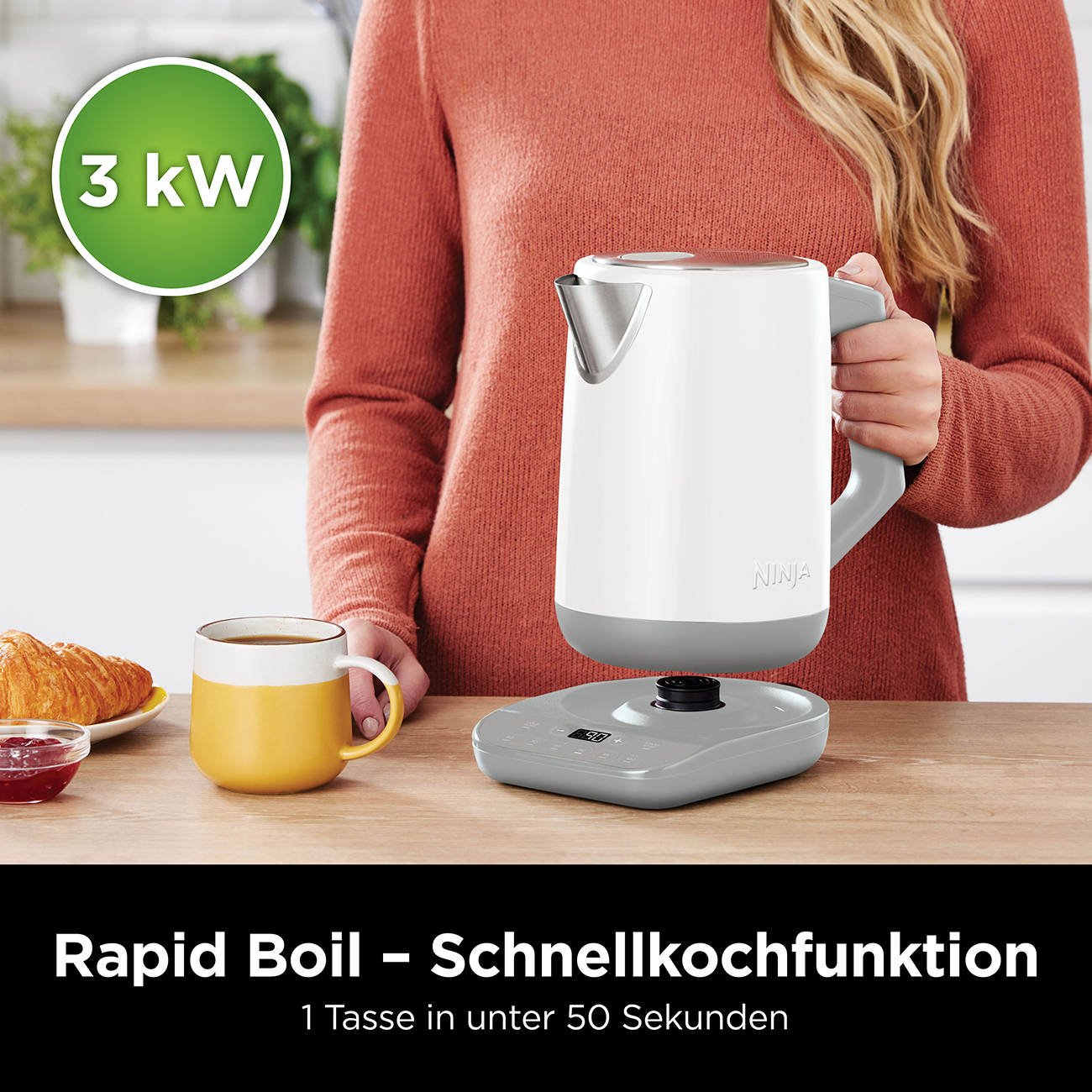 Donna che tiene un bollitore Ninja bianco. Una tazza con caffè e un croissant su un tavolo di legno.