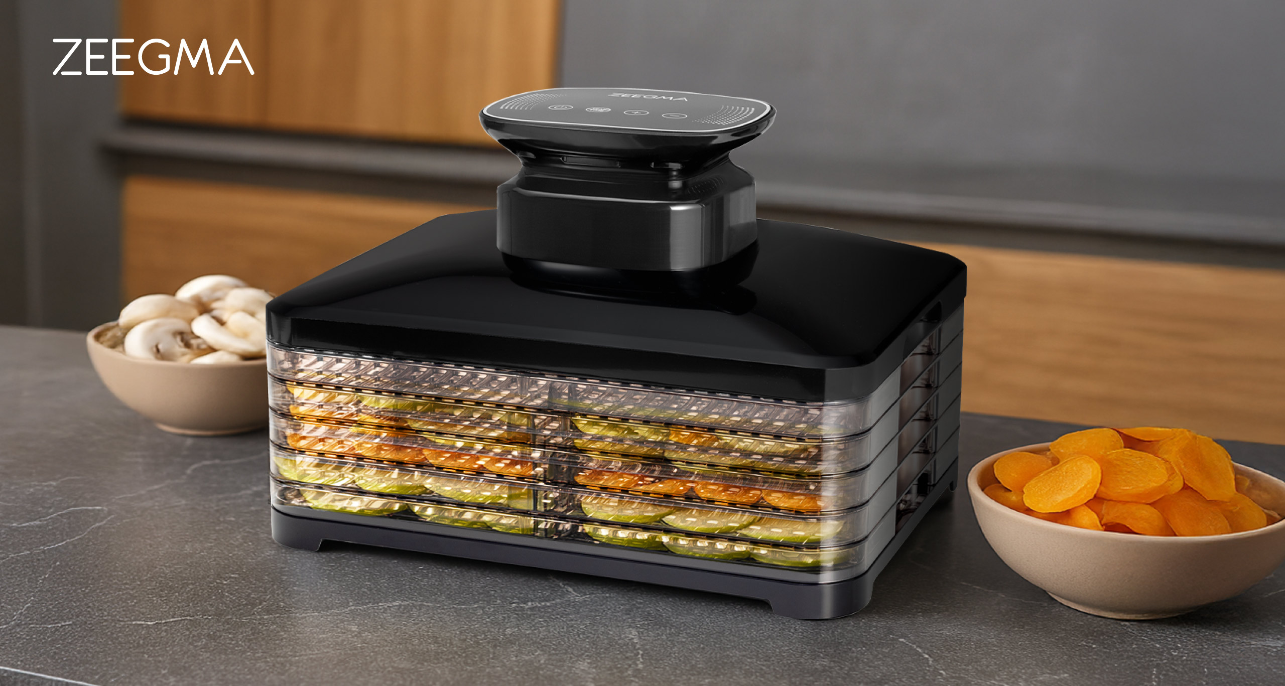 Czarny dehydrator do żywności z tacami z jedzeniem, miskami z pieczarkami i morelami na blacie kuchennym.
