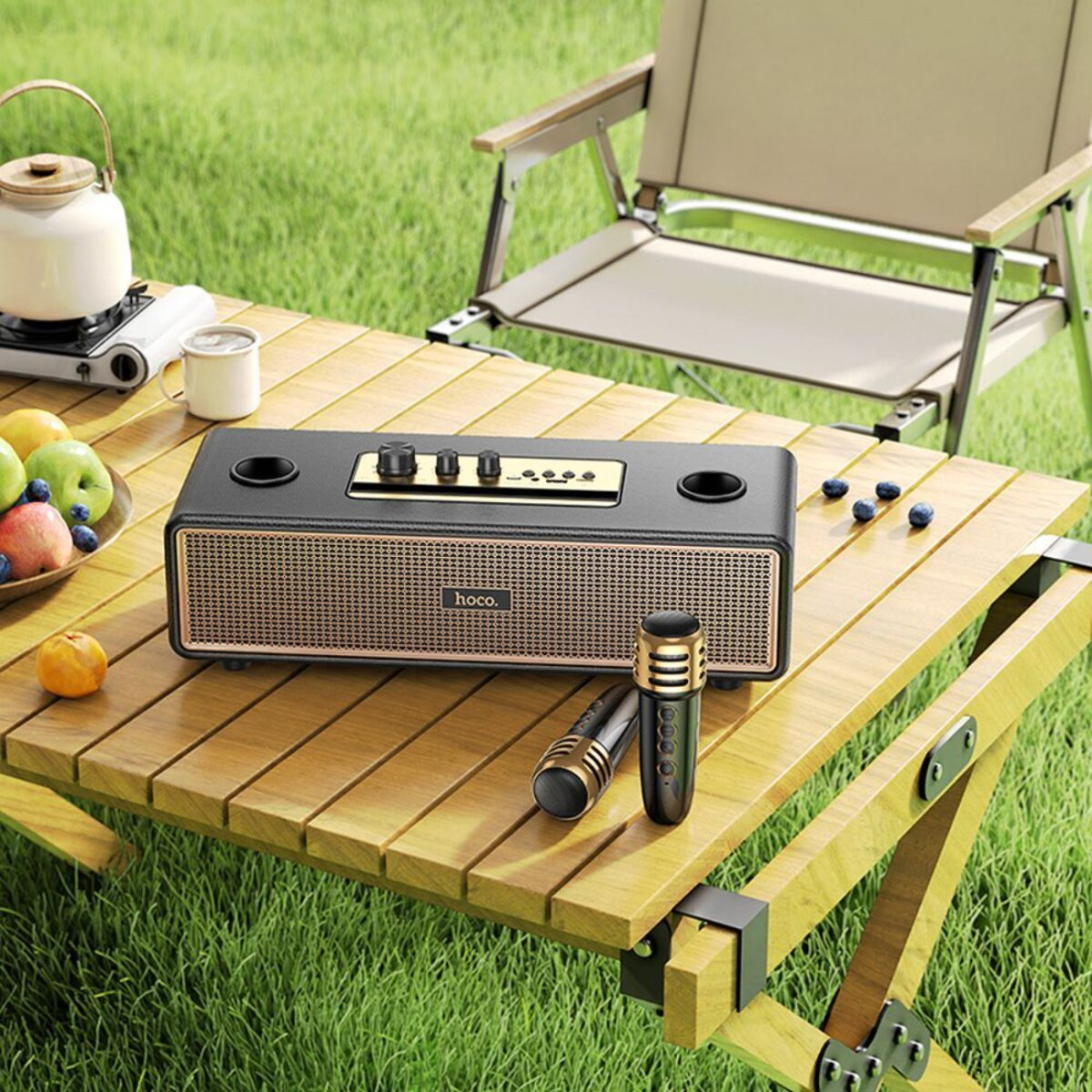 Un altoparlante poggia su un tavolo di legno con un'ambientazione da picnic. Include un bollitore, frutta e un microfono.