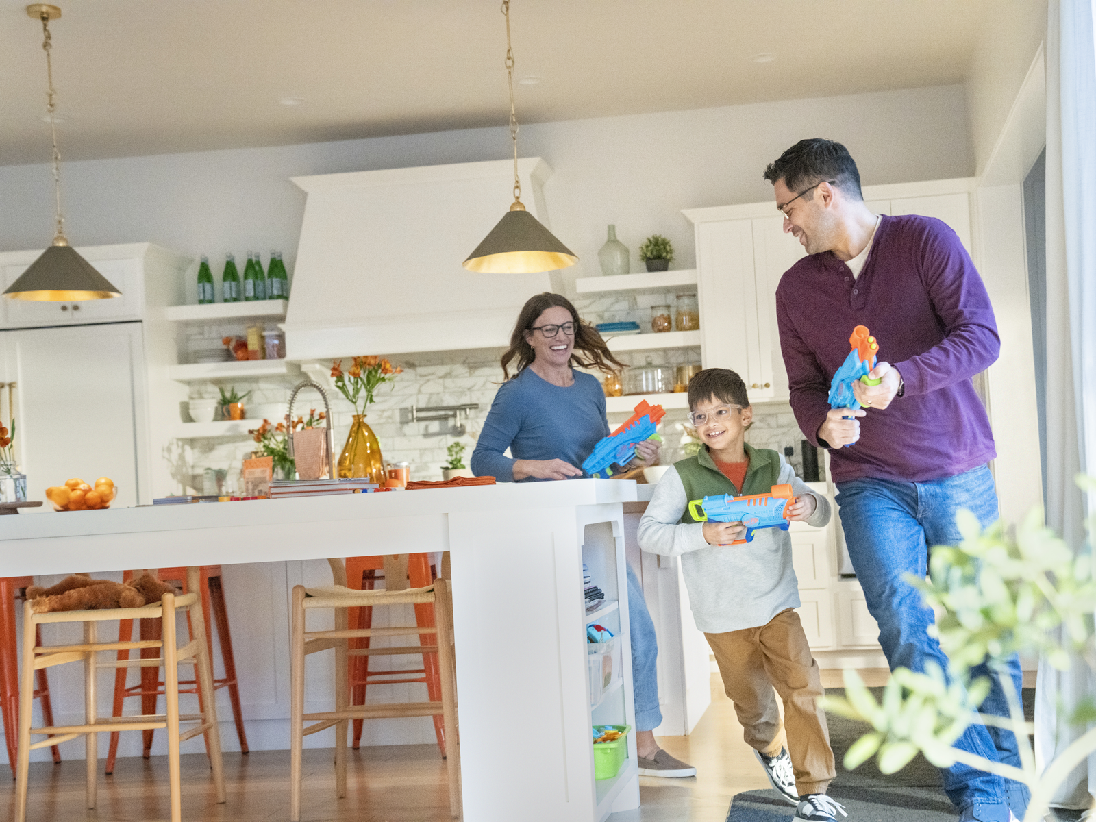 Una famiglia gioca con pistole ad acqua in una cucina luminosa. Isola bianca e pavimenti in legno visibili.