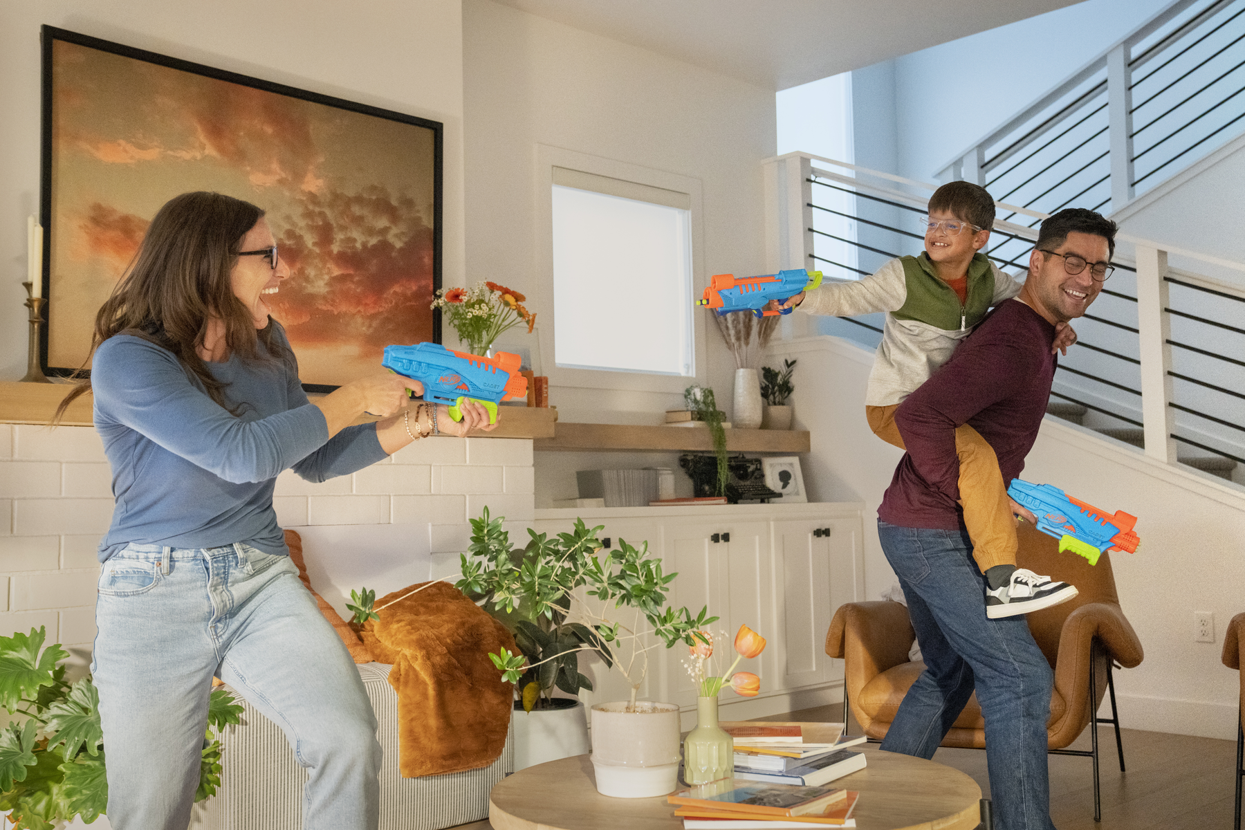 Famiglia che gioca con pistole ad acqua in casa. Madre, figlio sulla schiena del padre, tutti sorridenti. Interno con arte e piante.