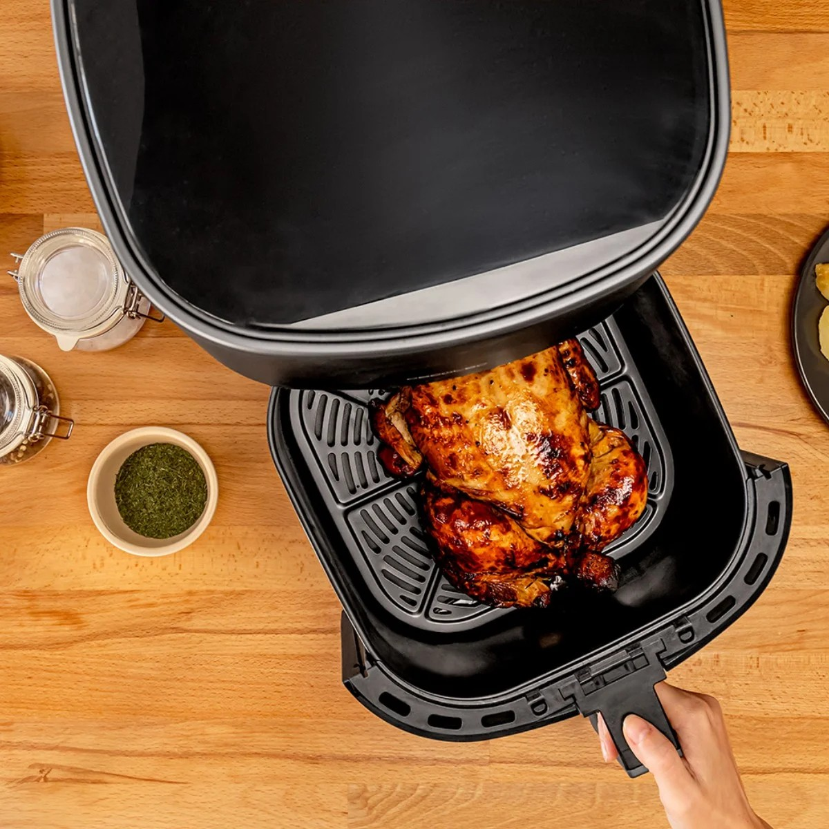 Vista cenital de freidora con pollo asado dentro. Especias en cuencos. Mesa de madera.
