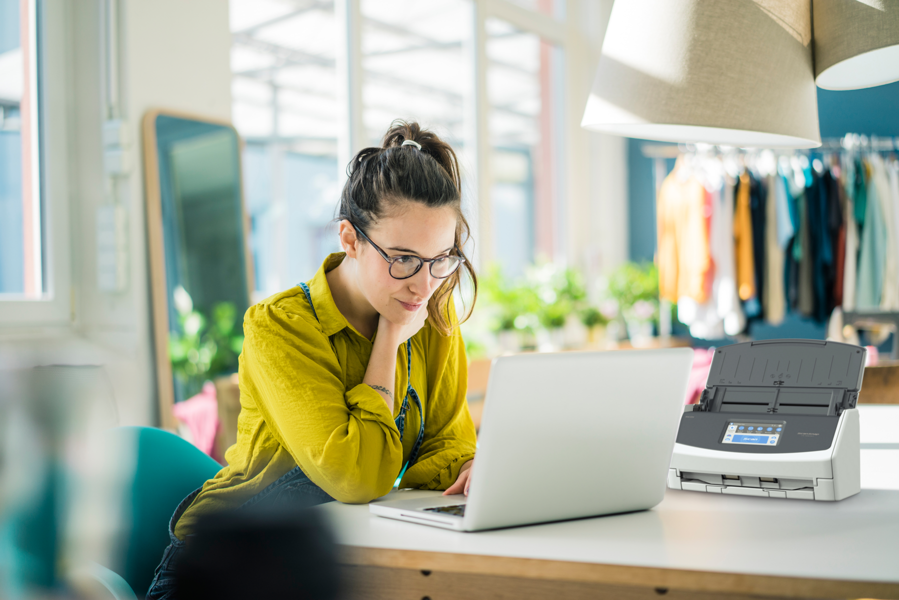 Kobieta używa laptopa przy biurku z drukarką. Pokój ma naturalne światło.