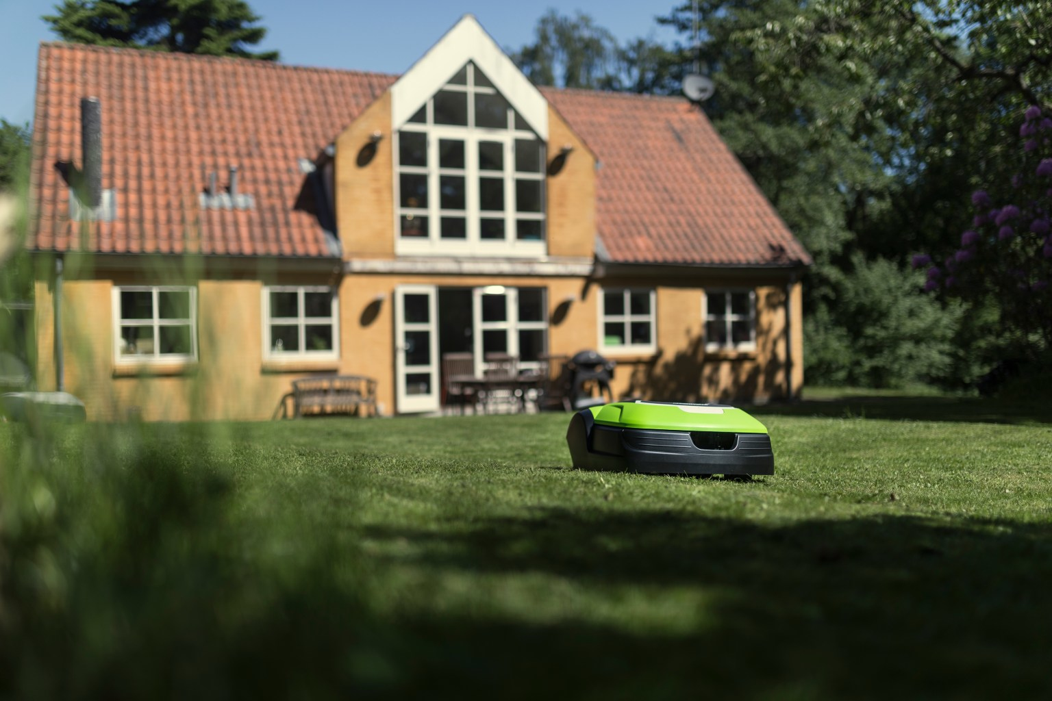 Ein grüner und schwarzer Mähroboter auf einem Rasen vor einem gelben Haus mit rotem Dach.