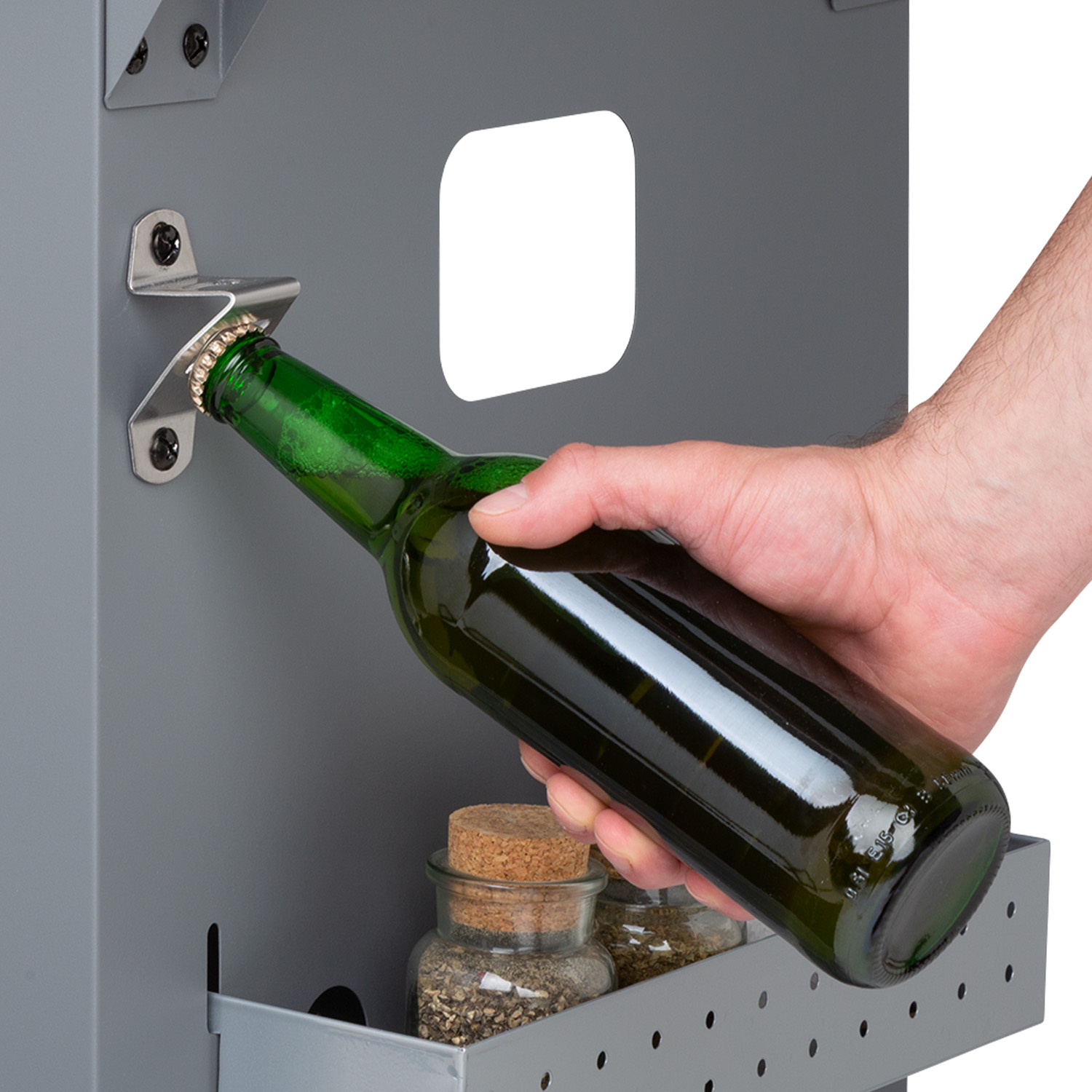 Eine Hand benutzt einen Flaschenöffner an einem Grill, um eine grüne Flasche zu öffnen, darunter stehen Gewürzgläser.