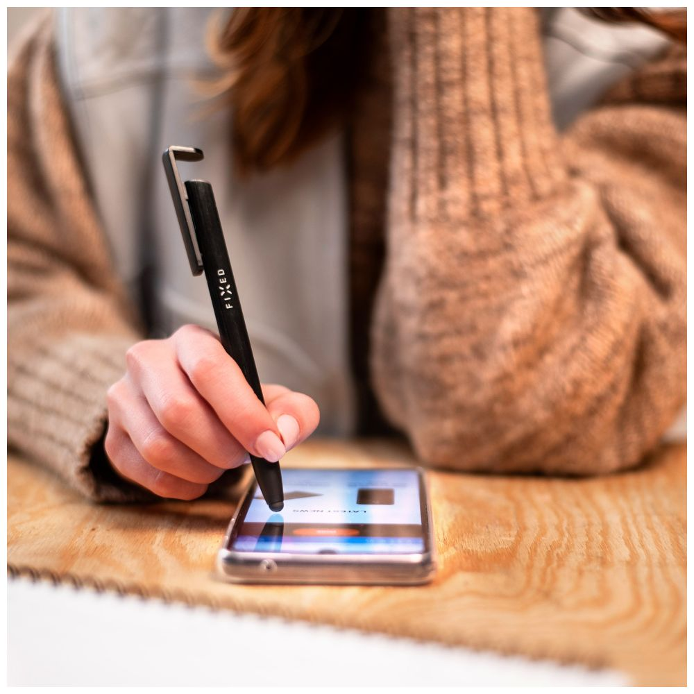 Die Hand einer Person hält einen Stift, um einen Telefonbildschirm auf einem Holztisch zu berühren.