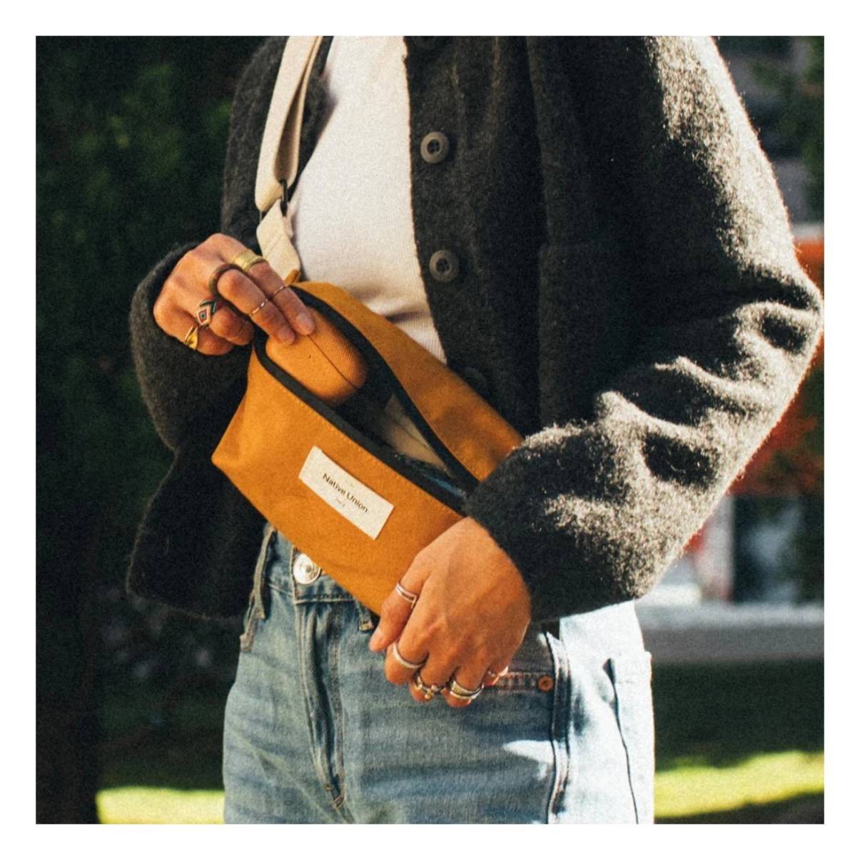 Una persona tiene in mano un marsupio giallo senape. Indossa una giacca grigia e jeans.