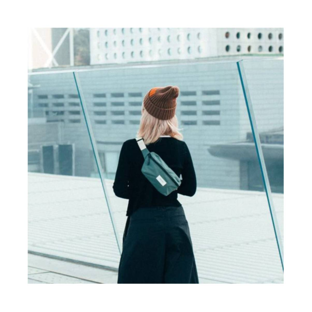Donna di spalle con borsa teal. Indossa un cappello e si trova vicino a finestre di vetro.
