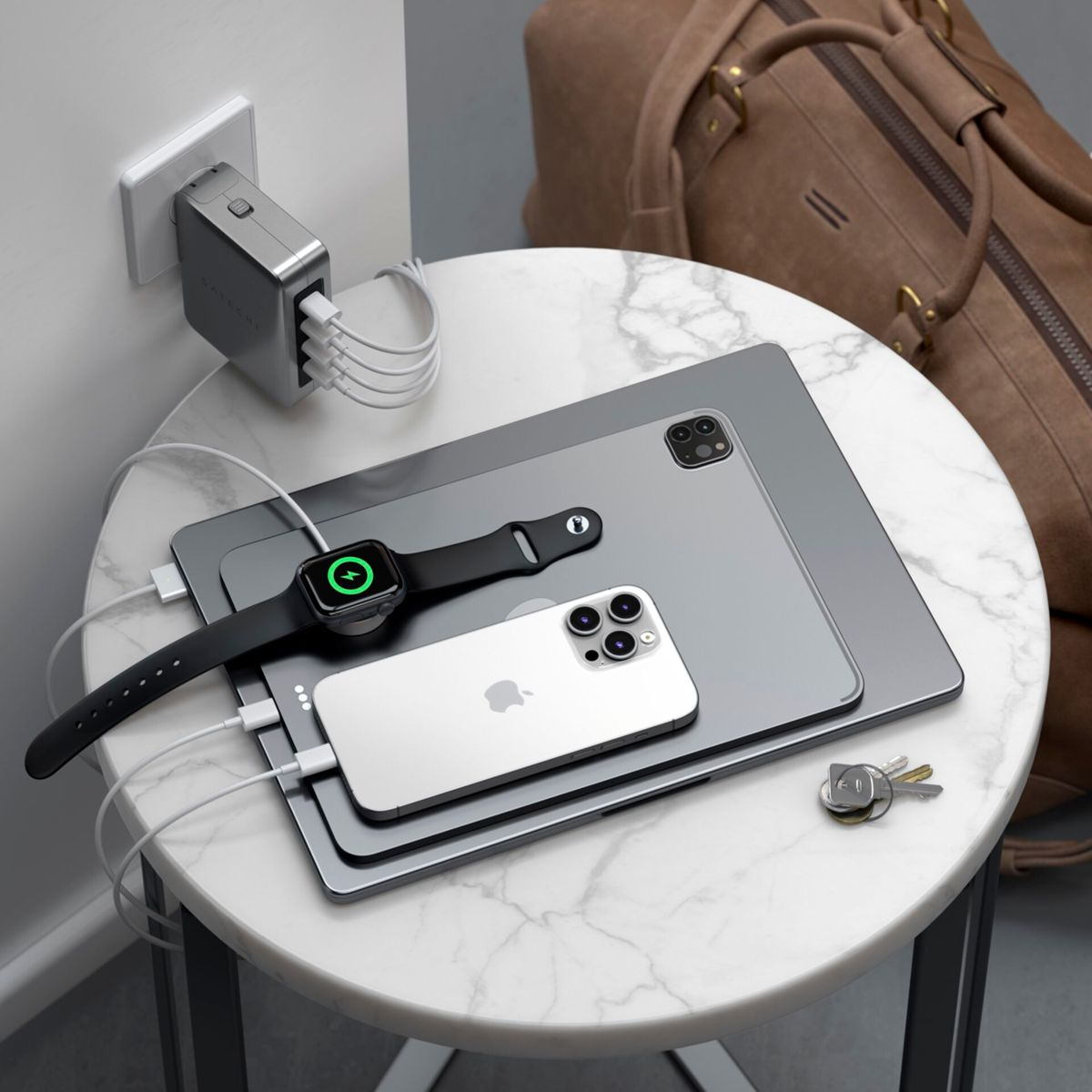 Più dispositivi in carica su un tavolo: iPhone, Apple Watch, iPad e laptop. Una borsa marrone dietro.