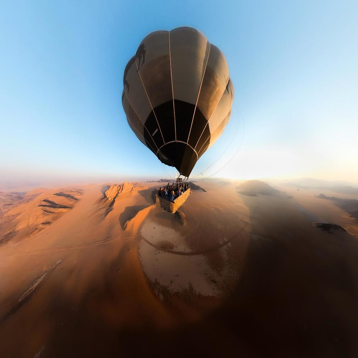 Heißluftballon über Wüste. Passagiere im Korb. Blauer Himmel, sandiges Gelände und ferne Berge.