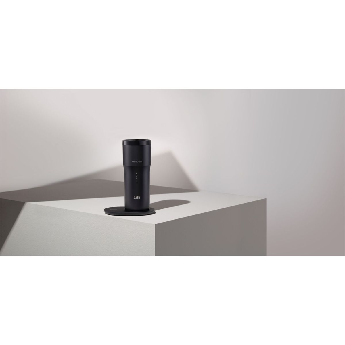 Una tazza Ember nera con base nera, che mostra la temperatura 135 su un cubo bianco.