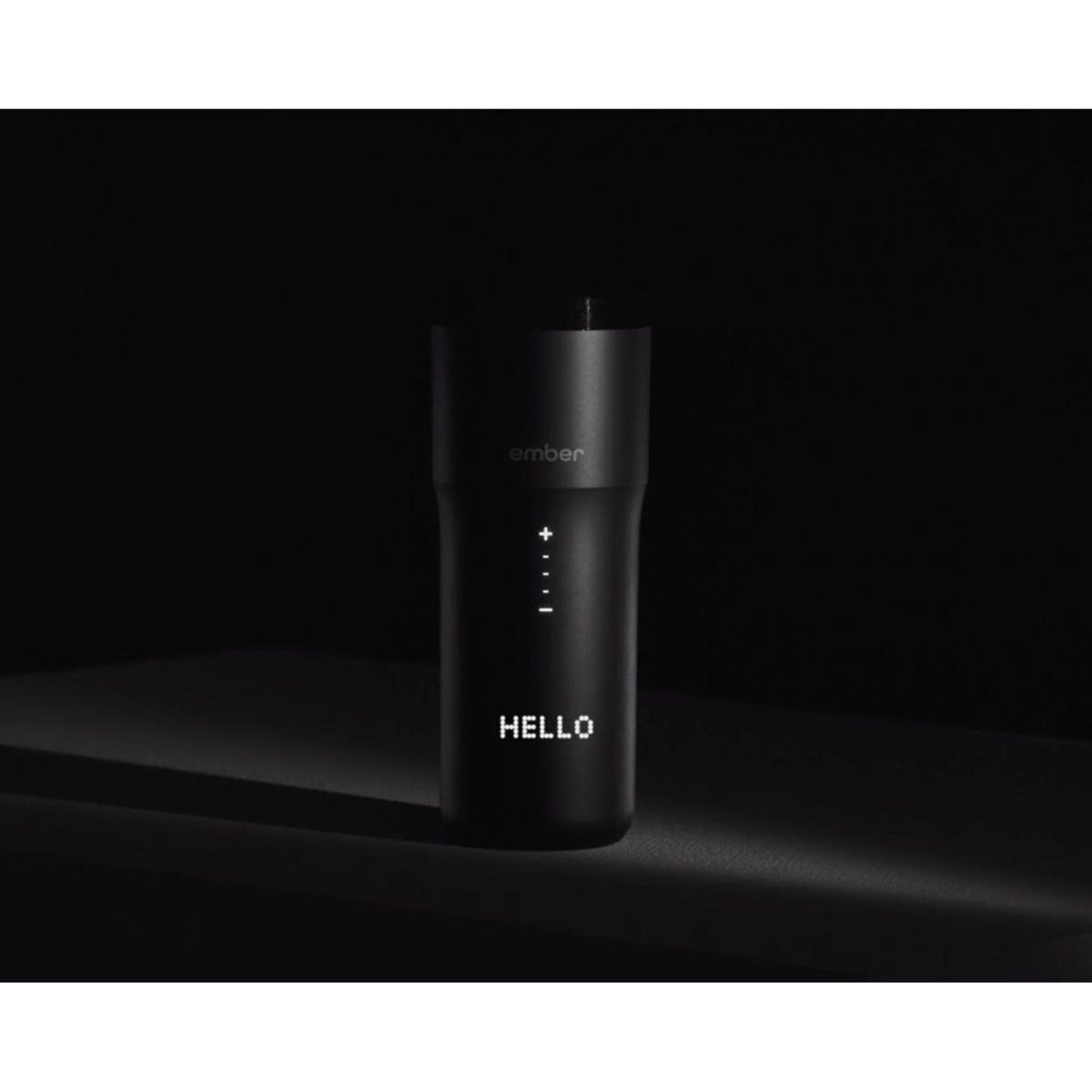 Tazza da viaggio Ember nera con display illuminato che mostra 'HELLO'. Sfondo e superficie scuri.