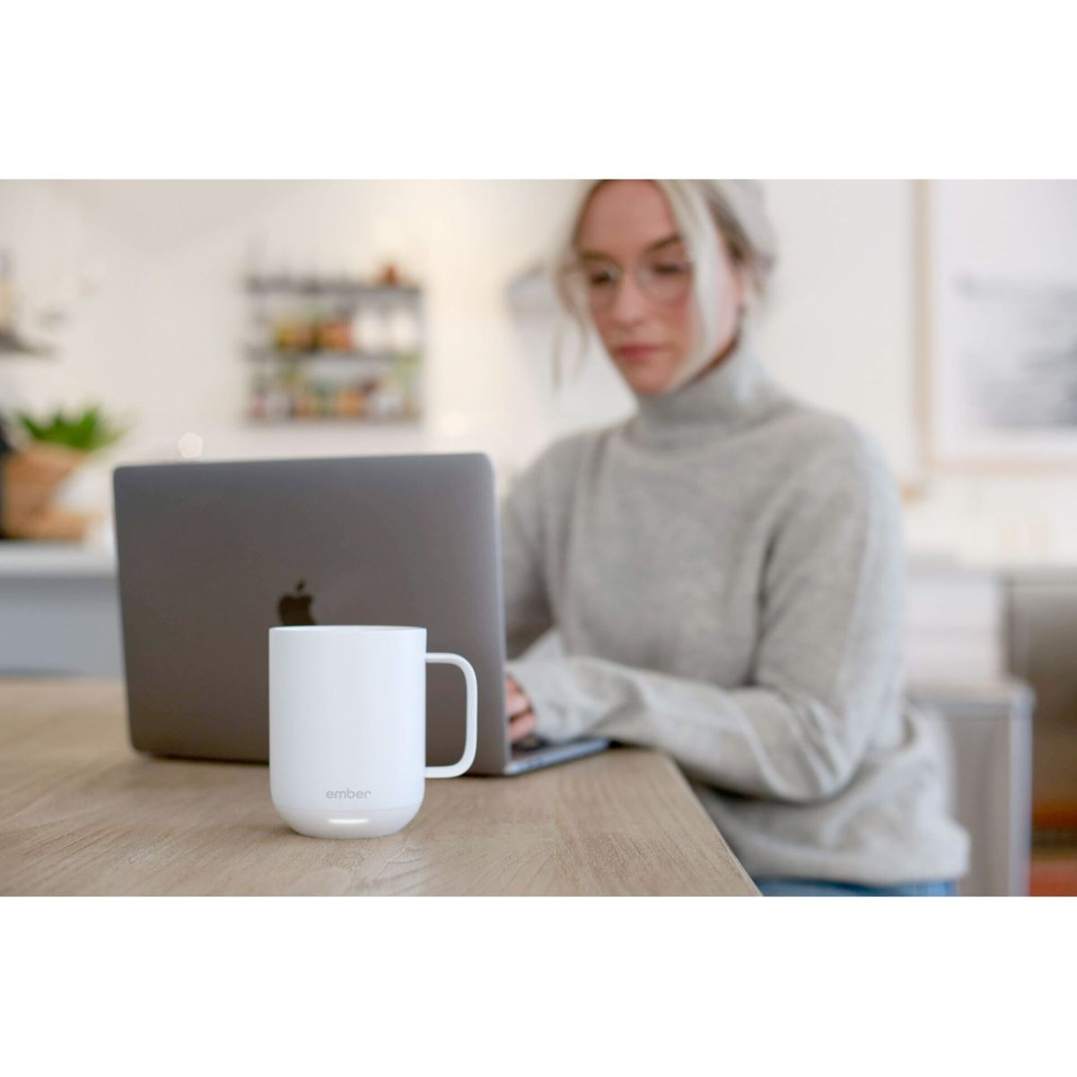 Donna con occhiali che usa un laptop, una tazza bianca sul tavolo. Sfondo cucina.
