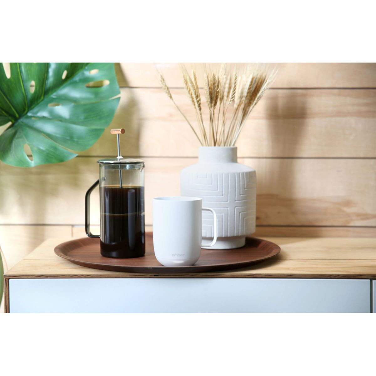 Tazza bianca su un vassoio di legno con pressa per caffè e vaso con spighe di grano. Mobile bianco sullo sfondo.