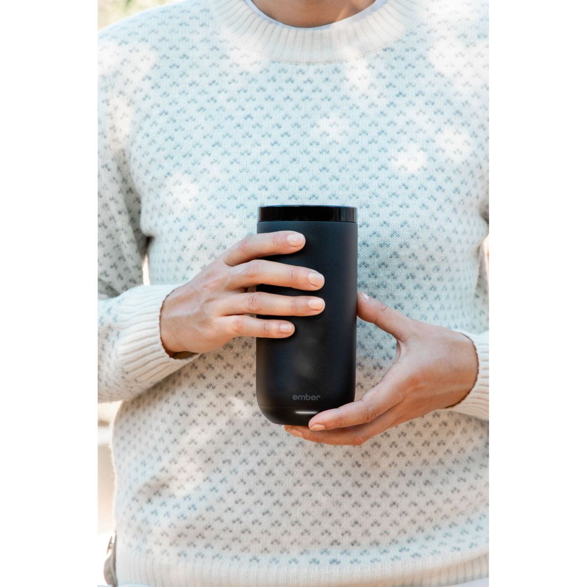 Eine Person hält eine schwarze, zylindrische Tasse. Die Tasse hat einen Henkel. Die Person trägt einen weißen Pullover.