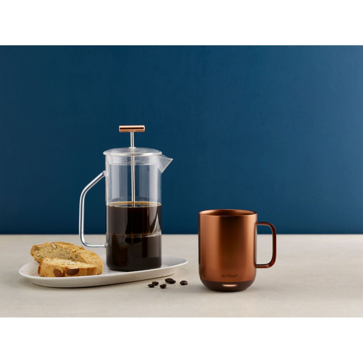 Una tazza di rame, una caffettiera a pistoni con caffè e biscotti su un vassoio bianco. Lo sfondo è un muro blu.