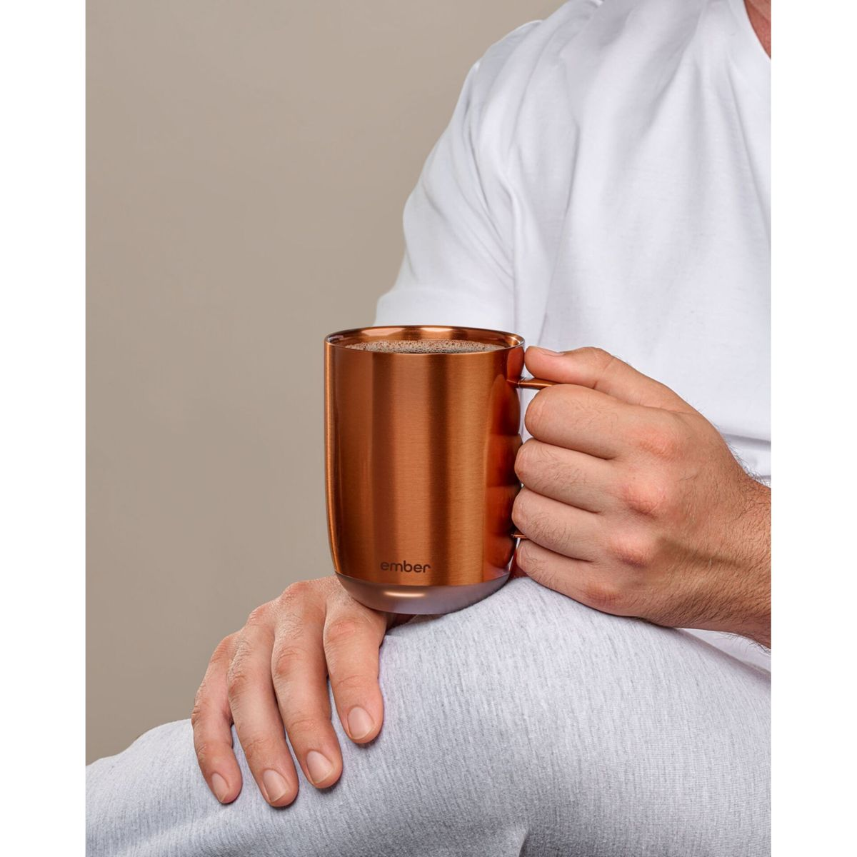 Una persona tiene una tazza di rame con manico, riempita con un liquido scuro. La persona indossa una camicia bianca e pantaloni grigi.