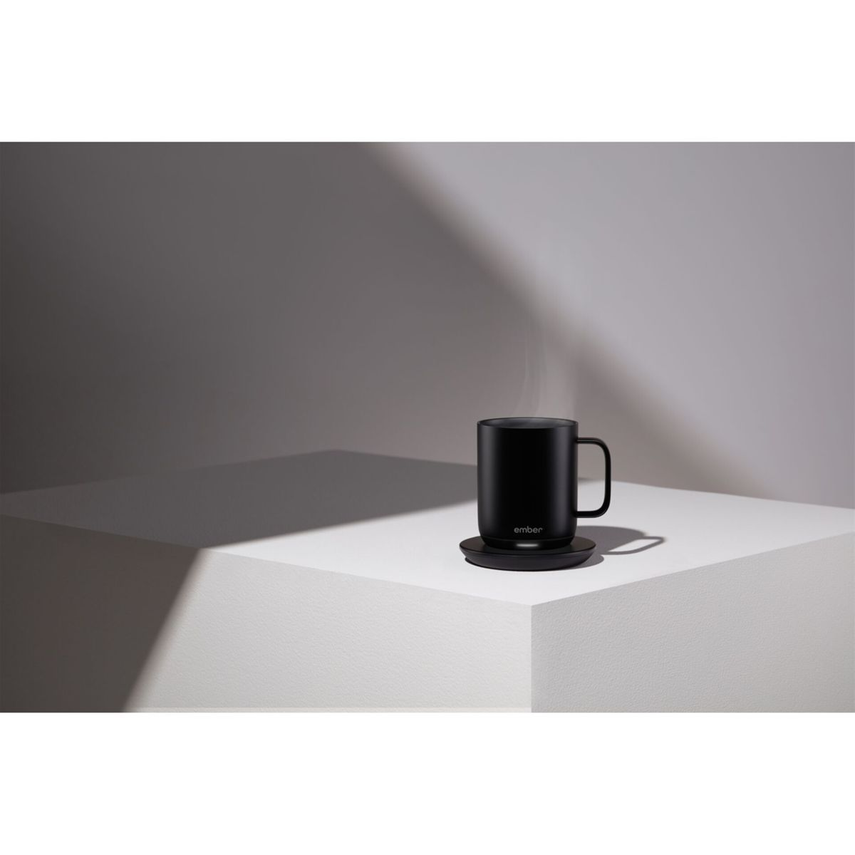 Una tazza nera è su un sottobicchiere nero. Il vapore sale dalla tazza. Lo sfondo è bianco e grigio.