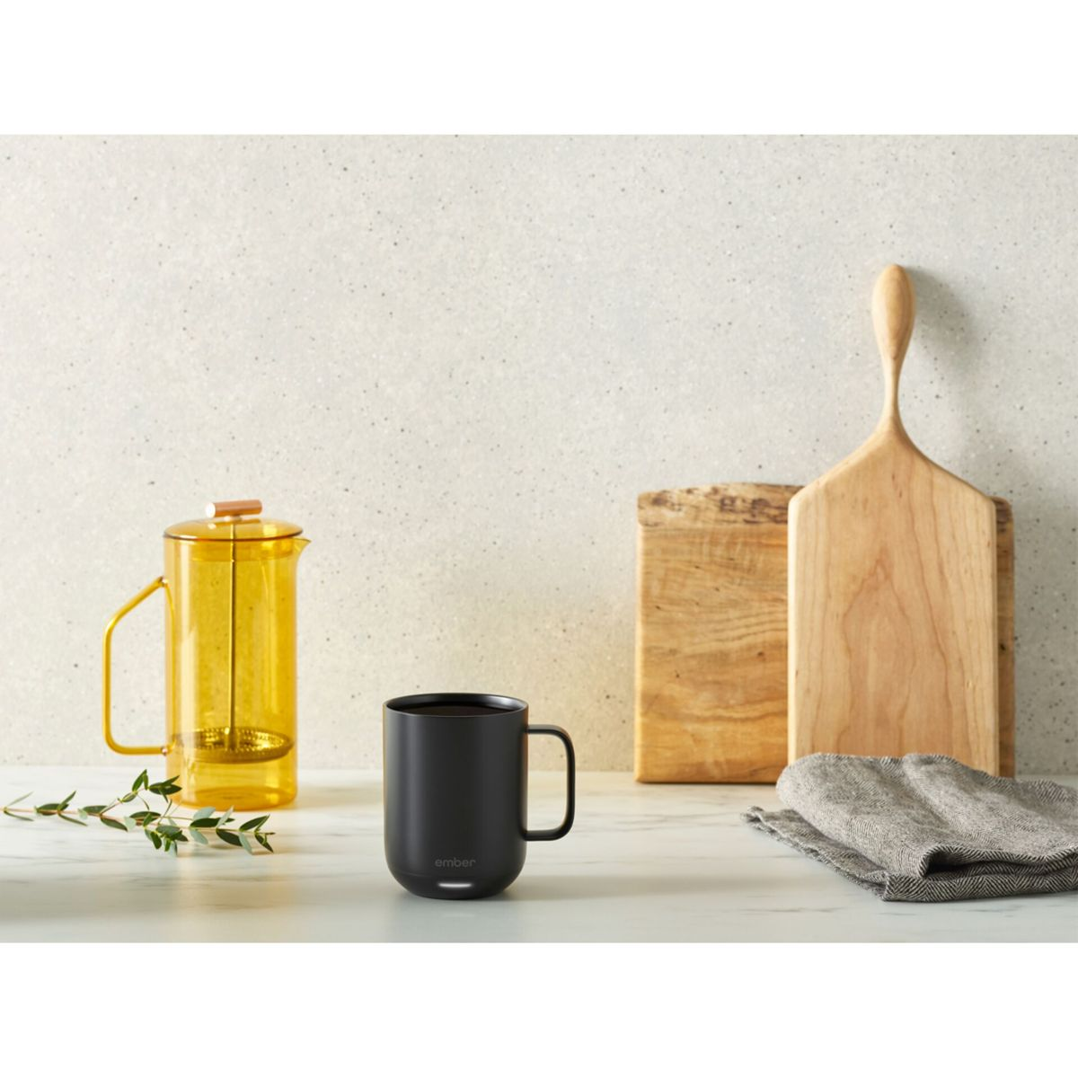 Una tazza nera, una caffettiera a pistoni gialla, un tagliere in legno e un panno grigio su un piano di lavoro.
