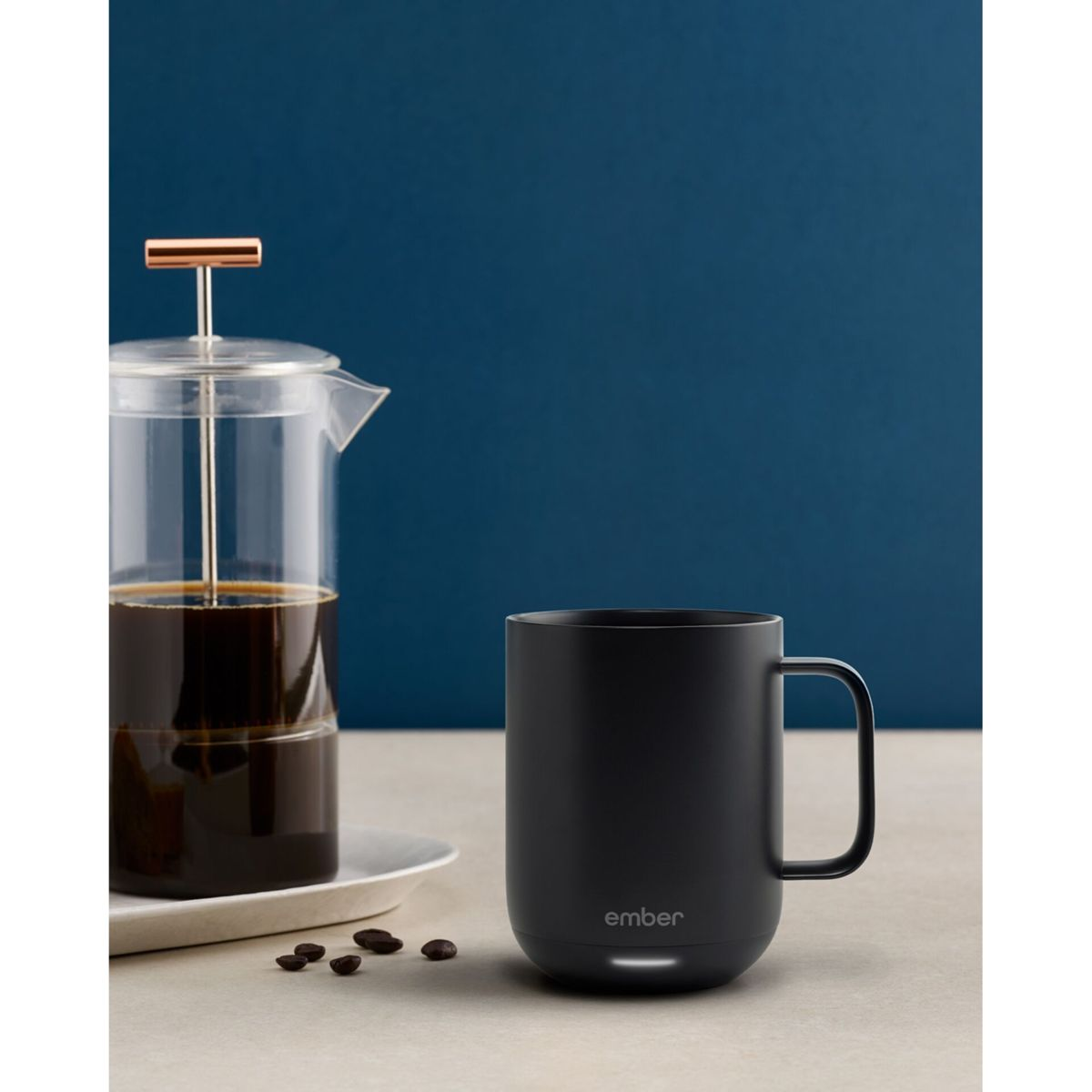 Una tazza nera, una caffettiera a pistoni con caffè e chicchi di caffè su una superficie bianca. Lo sfondo è un muro blu.