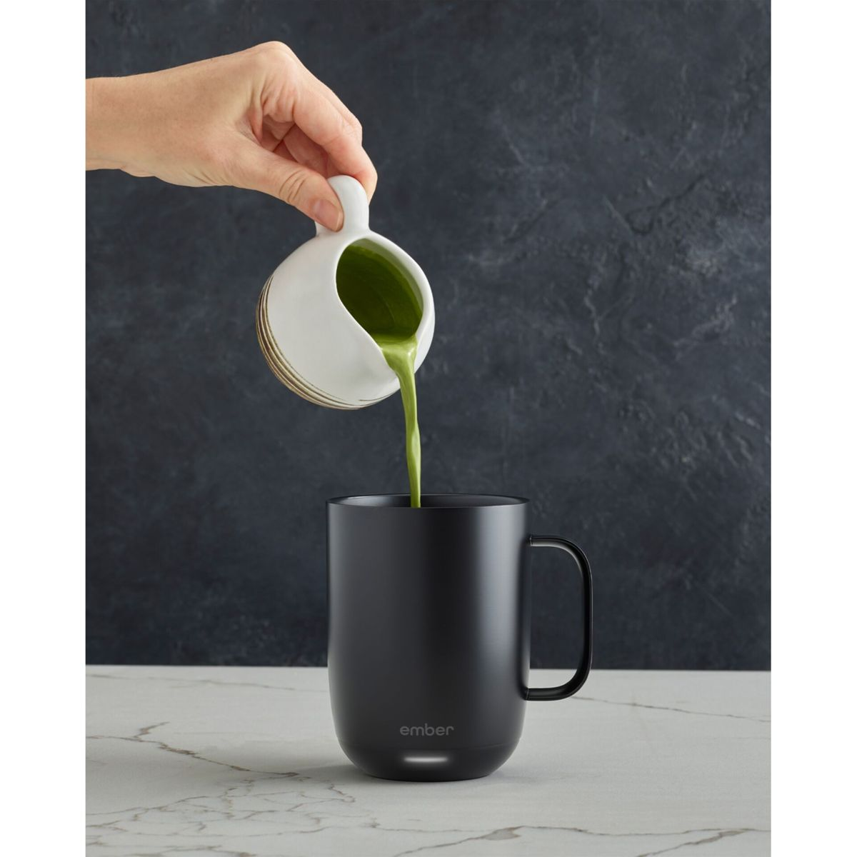 Una mano versa liquido verde da una lattiera bianca e dorata in una tazza nera. Lo sfondo è scuro.