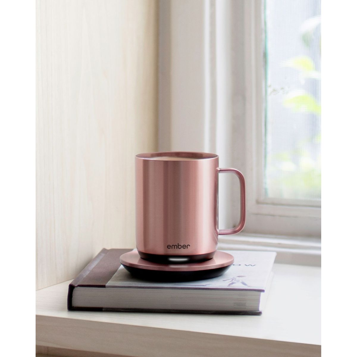 Tazza color oro rosa su un libro e un sottobicchiere. La tazza è vicino a una finestra. Lo sfondo è un muro di legno chiaro.
