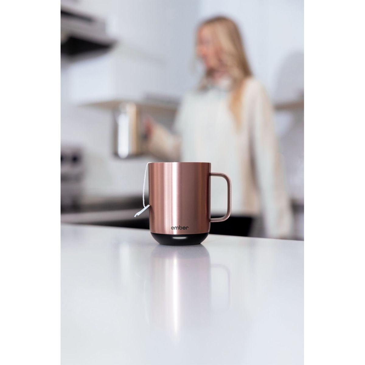Tazza color rame con base nera, appoggiata su una superficie bianca. Una donna sfocata sullo sfondo.
