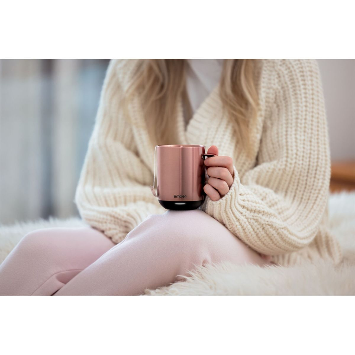 Donna seduta su un tappeto di pelliccia, che tiene in mano una tazza color rame. Indossa un maglione bianco e pantaloni rosa.