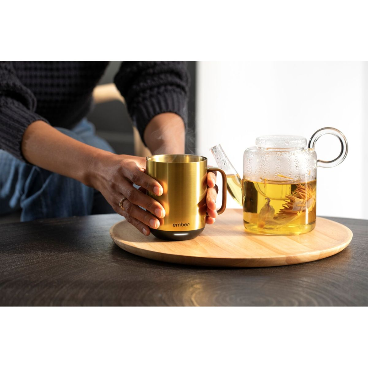 Hände halten einen goldenen Becher neben einer Glasteekanne. Ein Holztablett wird verwendet. Dampf ist sichtbar.