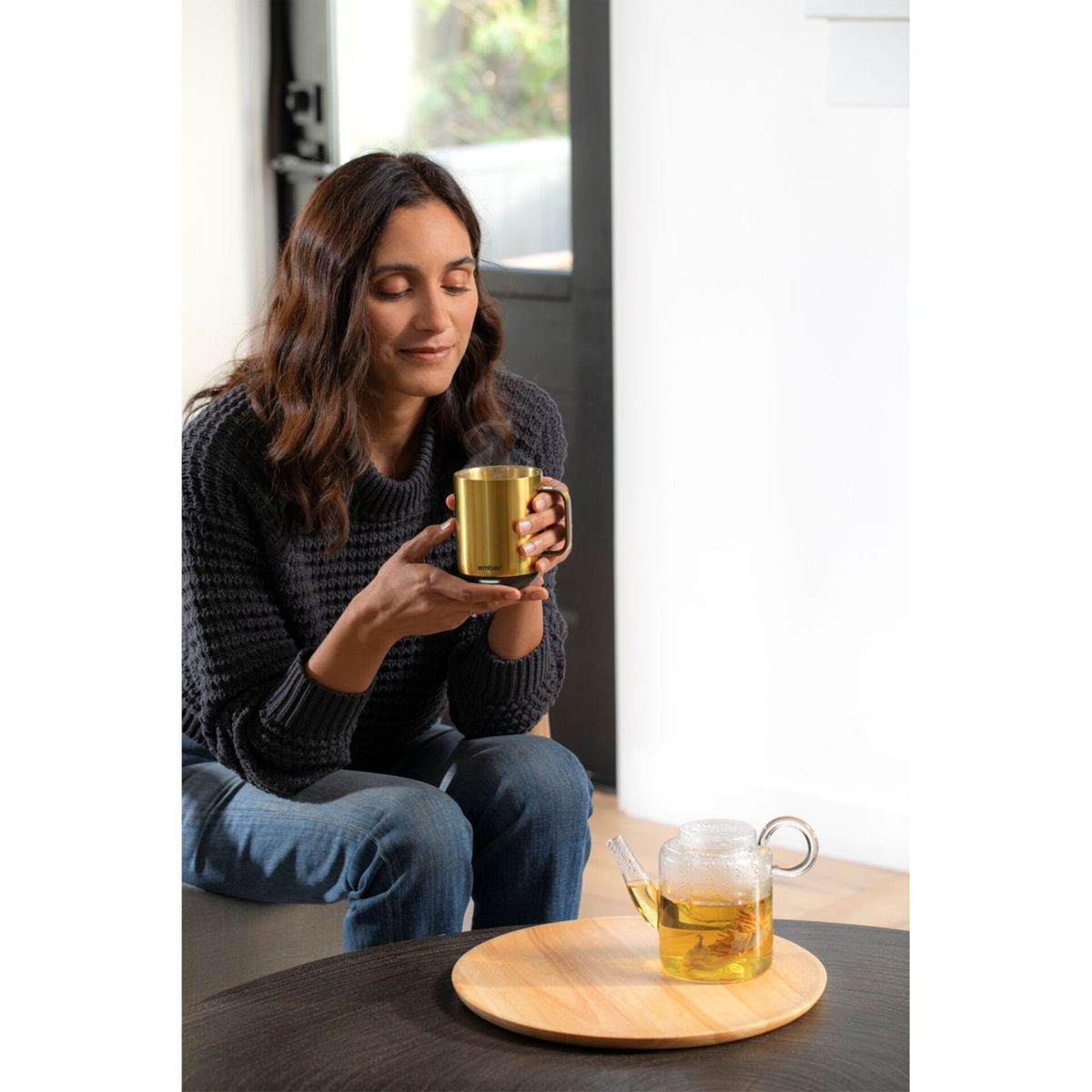 Frau hält einen goldenen Becher und trinkt Tee. Auf dem Tisch stehen eine Glasteekanne und ein Holztablett. Der Hintergrund ist unscharf.