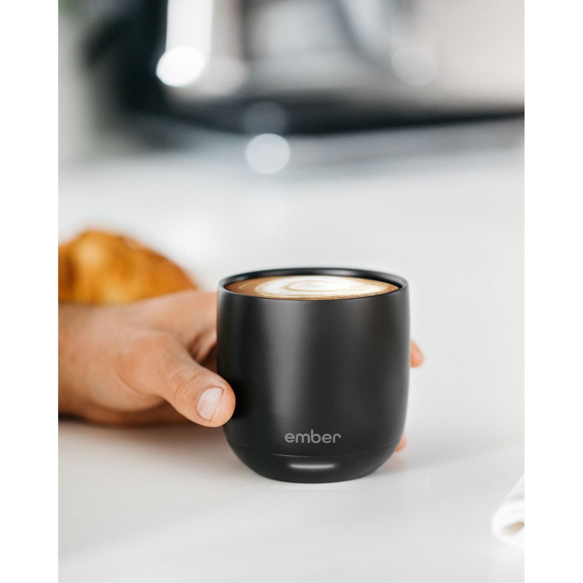 Una tazza nera con caffè è tenuta in mano. Un pasticcino sullo sfondo.