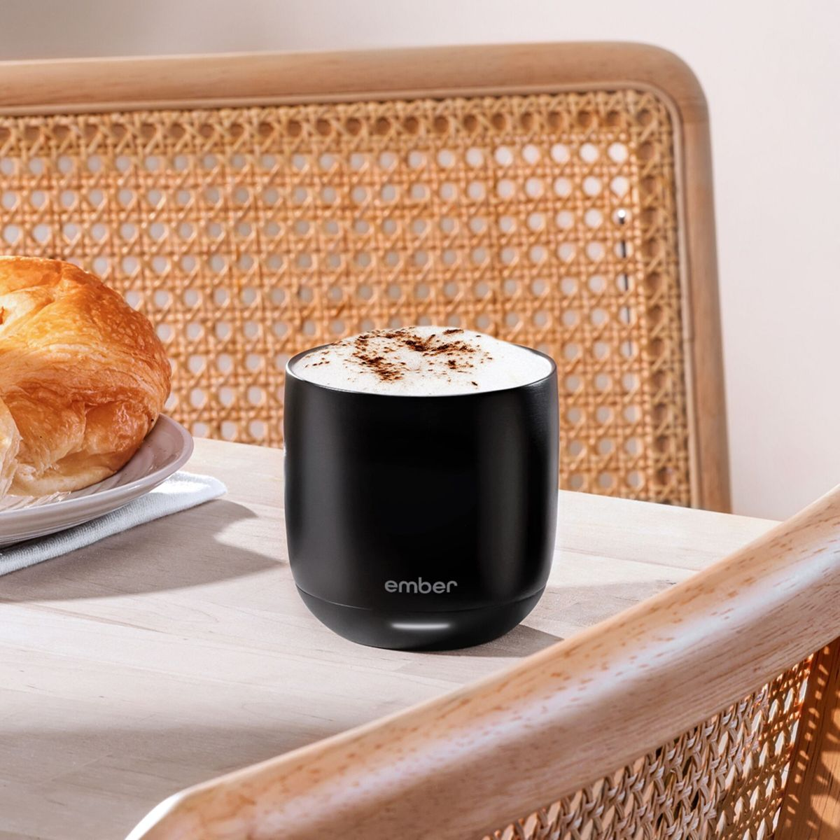 Una tazza nera con caffè su un tavolo. Un pasticcino e una sedia sullo sfondo.