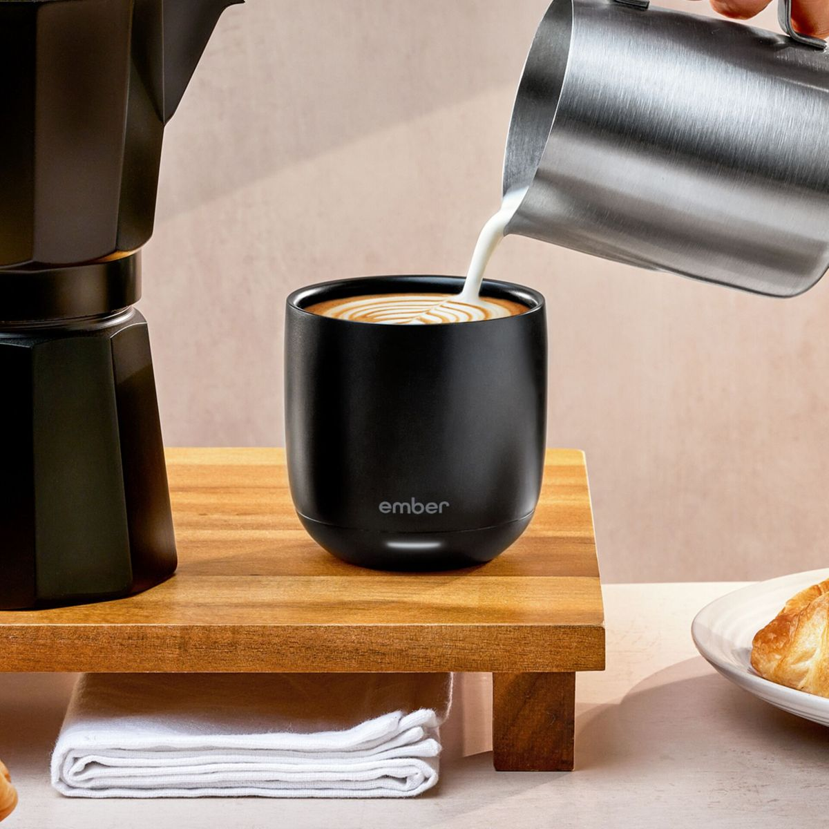 Latte versato in una tazza nera. La tazza è su una superficie di legno. Una macchina per il caffè e un pasticcino sono sullo sfondo.