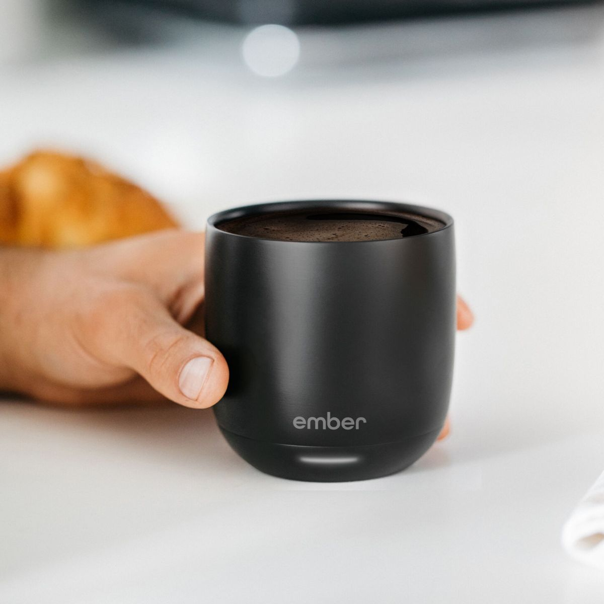 Una mano tiene una tazza nera con caffè. Un pasticcino sullo sfondo.