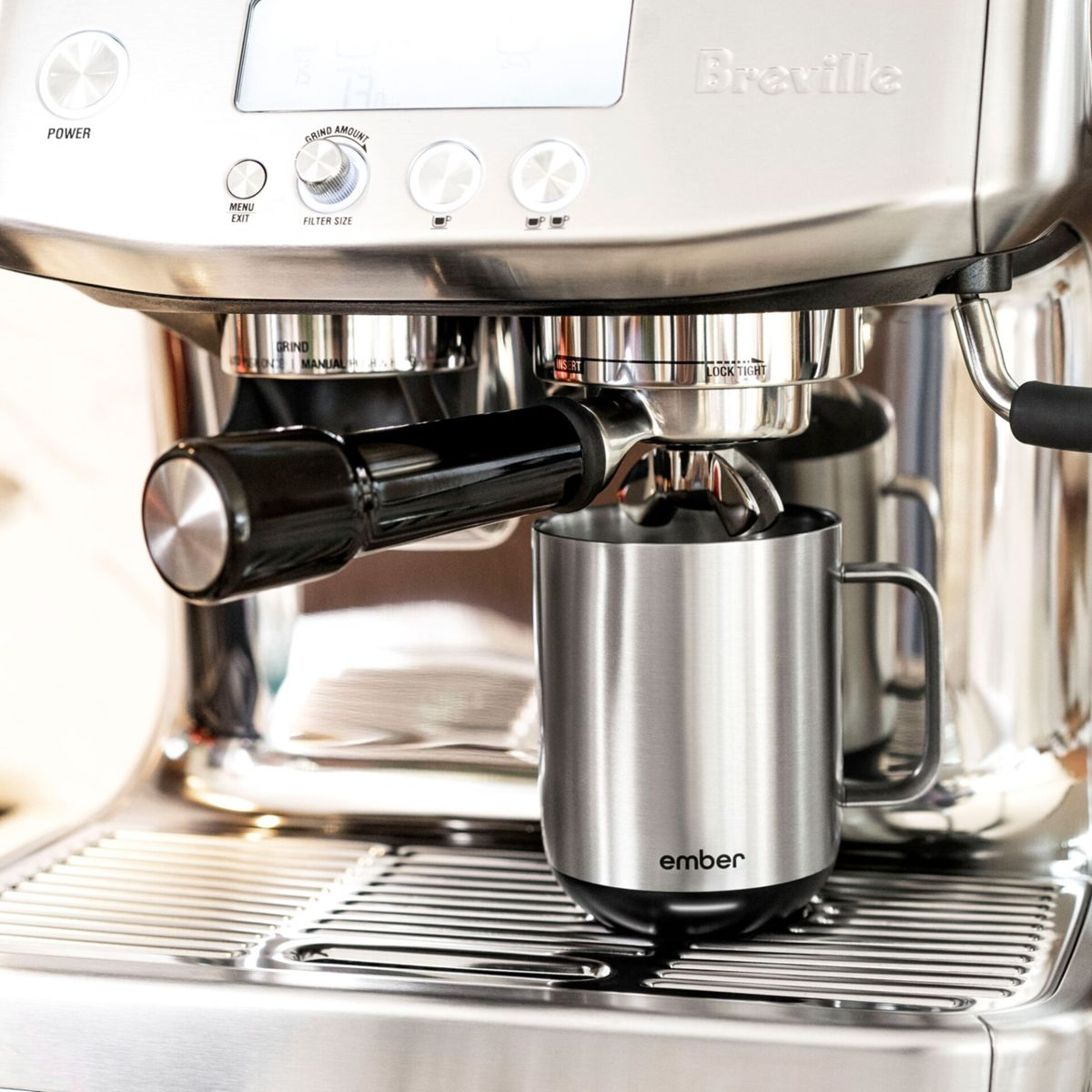 Una taza plateada en una máquina de espresso. La máquina tiene un mango negro y el logo Breville. El fondo está borroso.