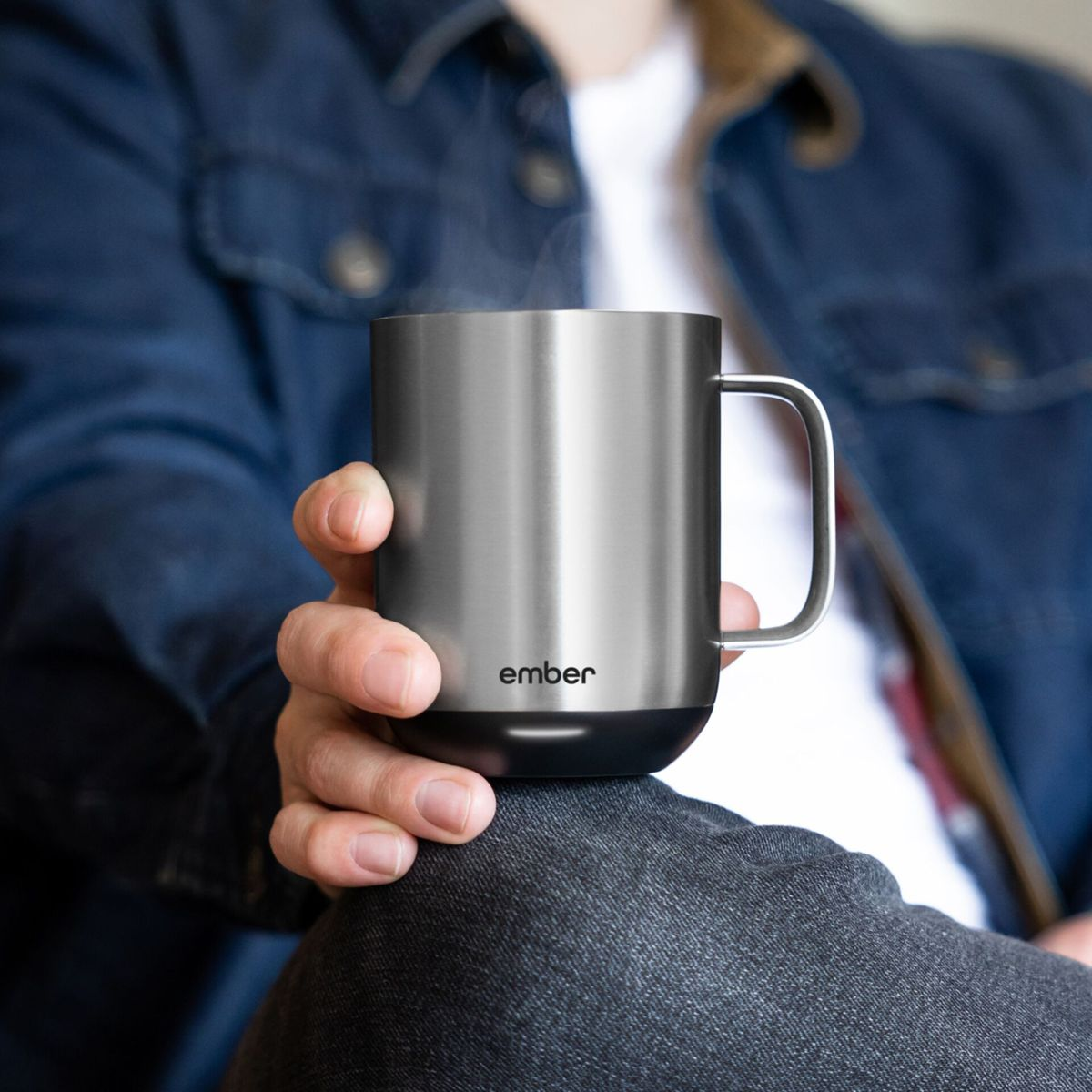 Un hombre sostiene una taza plateada. Lleva una chaqueta de mezclilla. Un fondo.