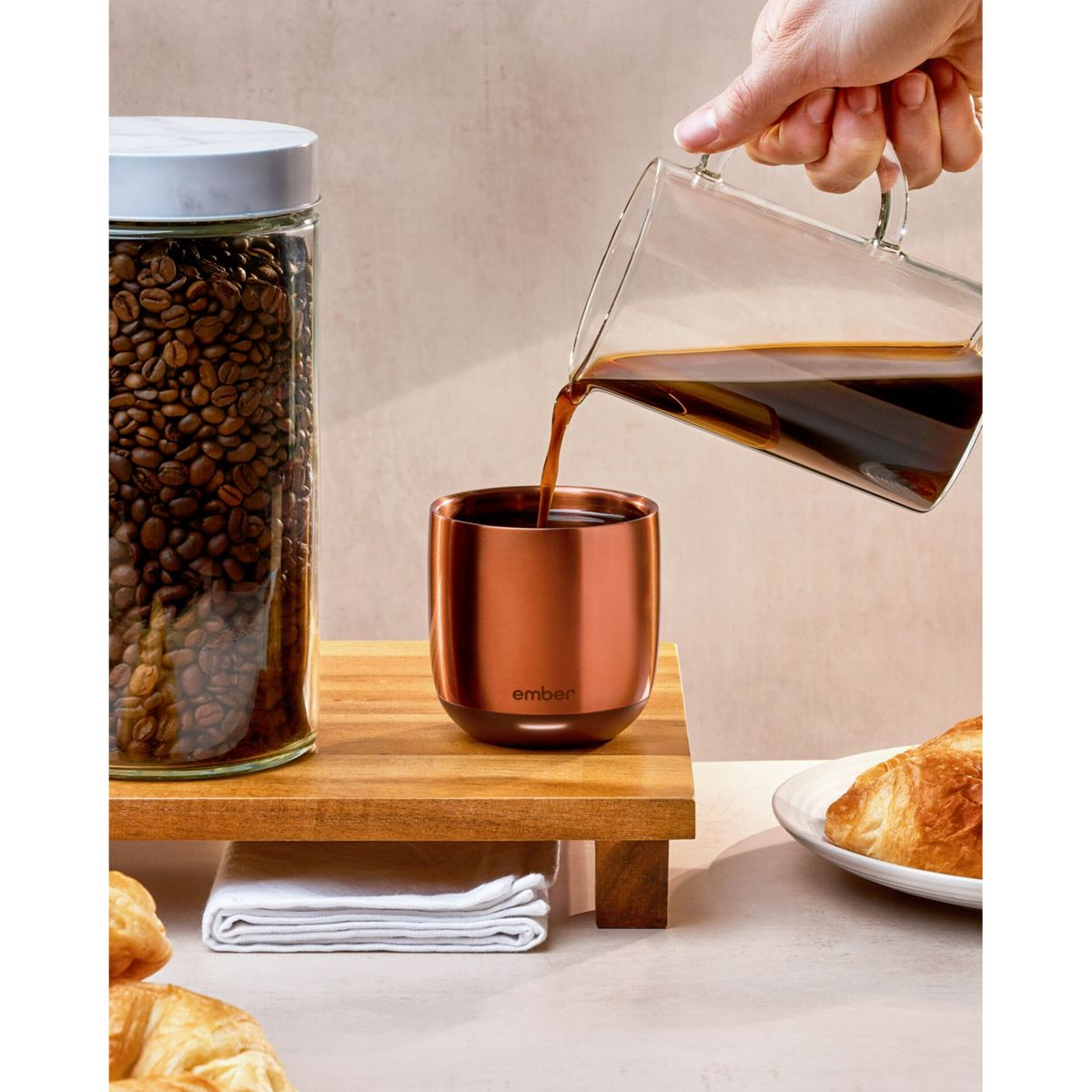 Caffè versato in una tazza di rame. Chicchi di caffè in un barattolo di vetro a sinistra e un croissant a destra.