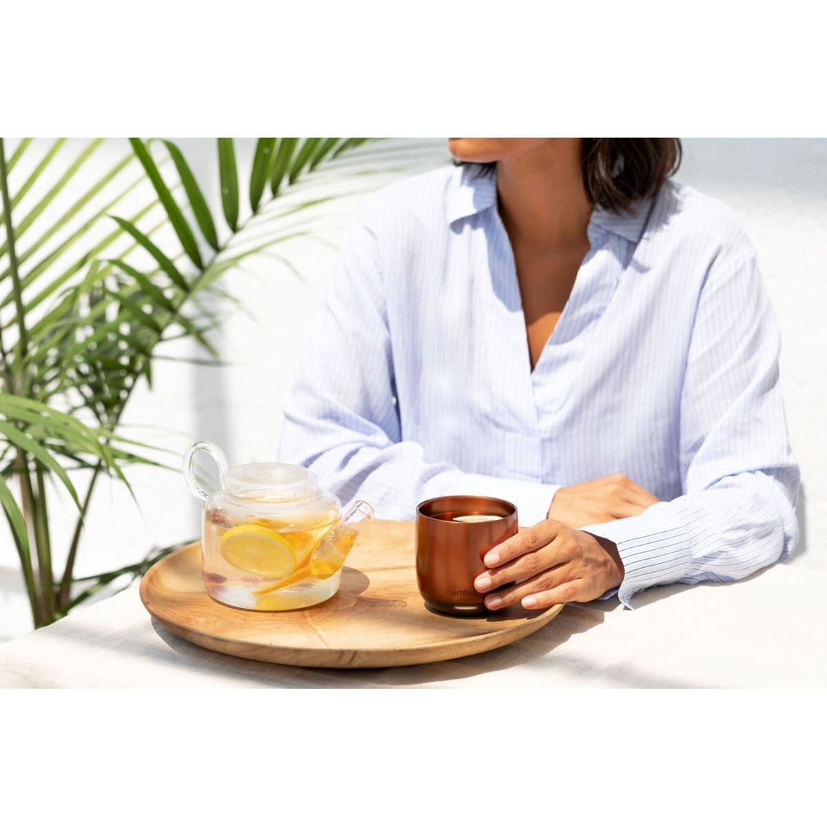 Donna seduta con tè e tazza su un vassoio di legno. Una teiera di vetro con fette di limone e foglie di palma sullo sfondo.