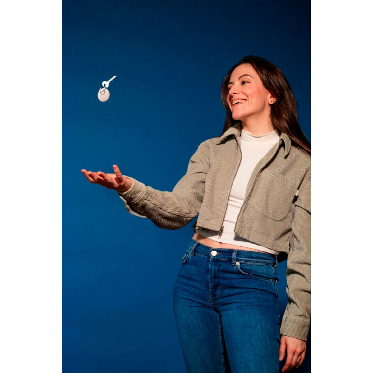 Kobieta rzuca w powietrze białą, okrągłą zawieszkę do kluczy. Uśmiecha się i ma niebieskie tło.