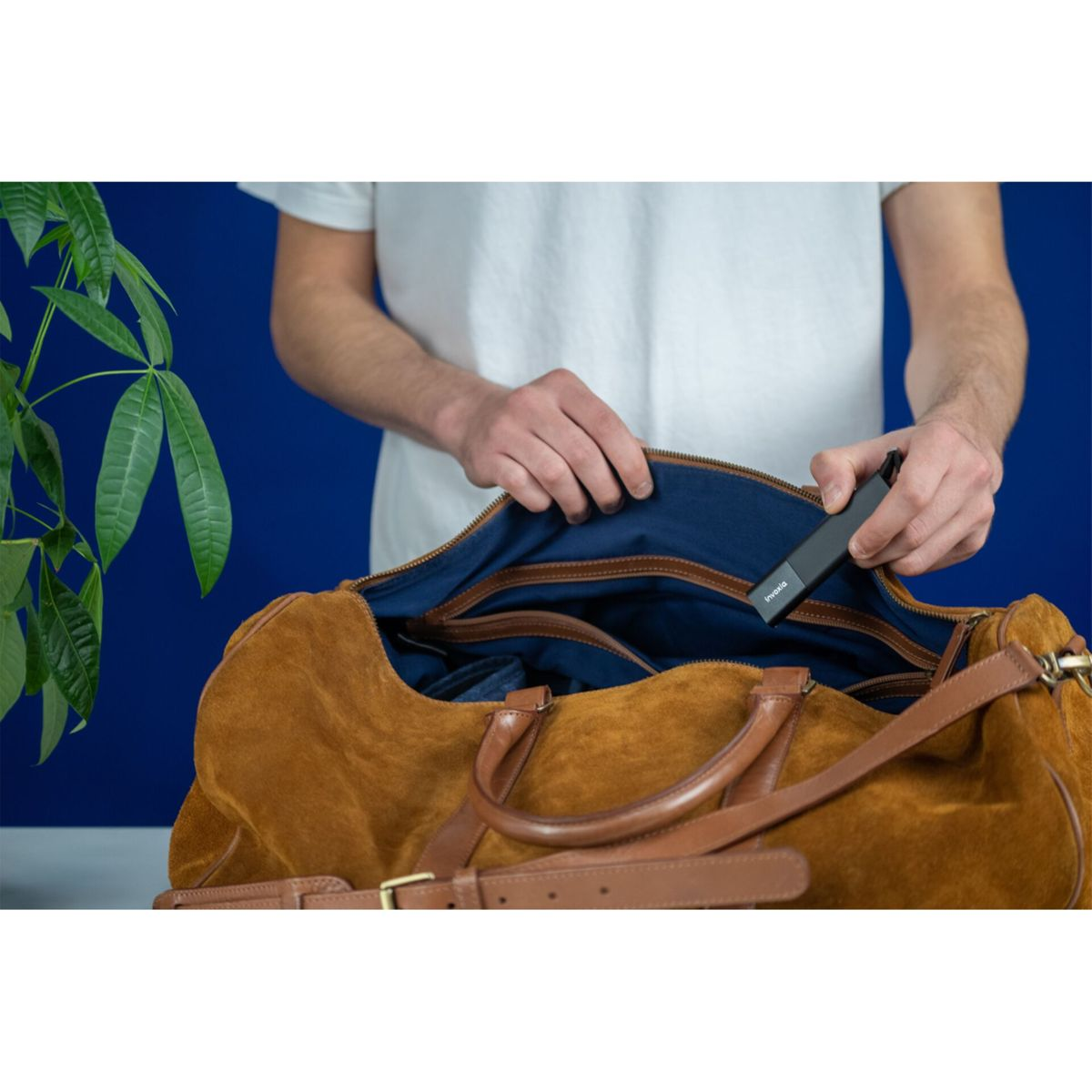 Osoba wkłada czarne urządzenie do brązowej torby. Tłem jest niebieska ściana i zielona roślina.