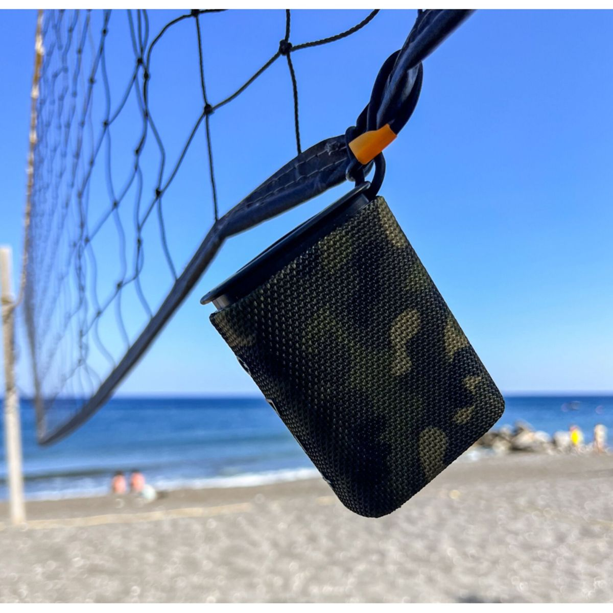 Una custodia mimetica è appesa a una rete da pallavolo. Lo sfondo è una spiaggia e cielo blu.
