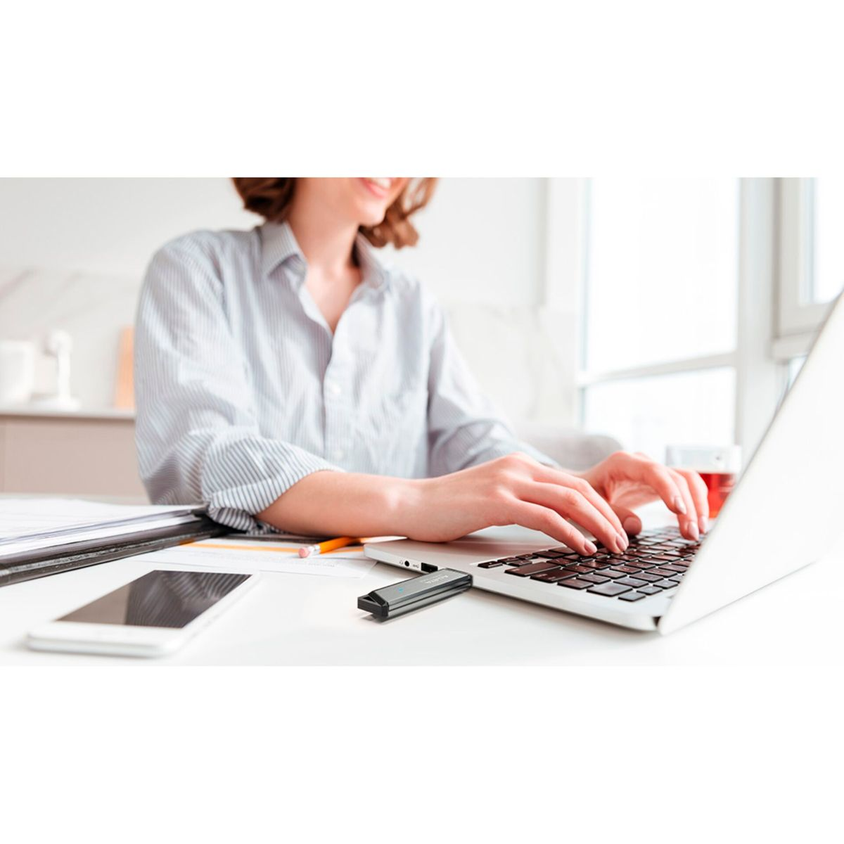 Eine Frau tippt auf einem Laptop, ein Telefon und ein USB-Laufwerk auf dem Tisch.