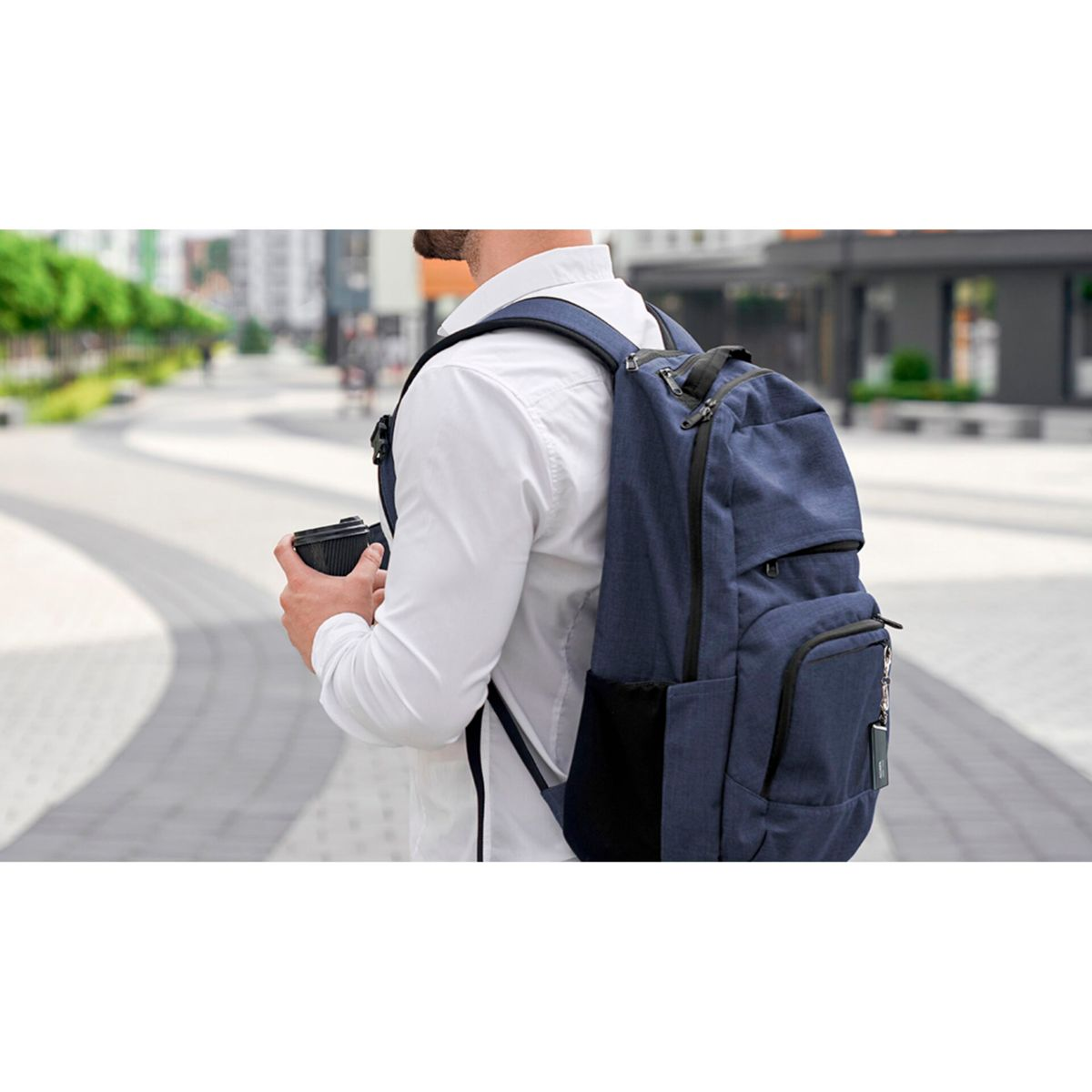 Ein Mann mit Rucksack und Kaffee geht eine Straße entlang. Gebäude sind im Hintergrund zu sehen.