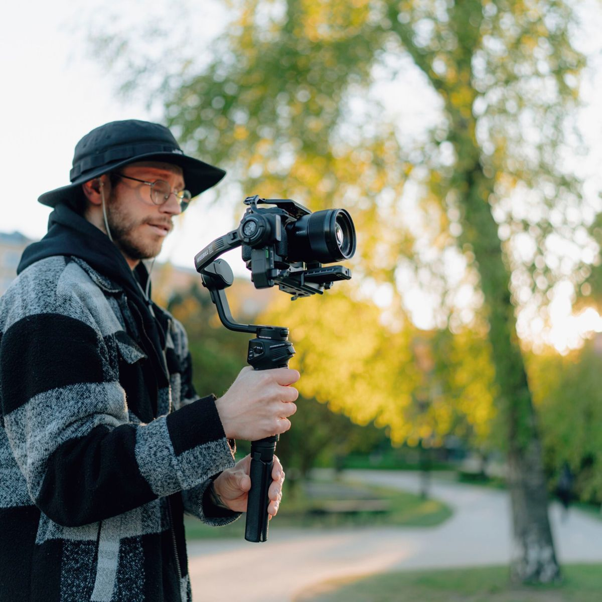 Mann mit Hut und Brille hält eine Kamera auf einem Gimbal, draußen mit Bäumen im Hintergrund.
