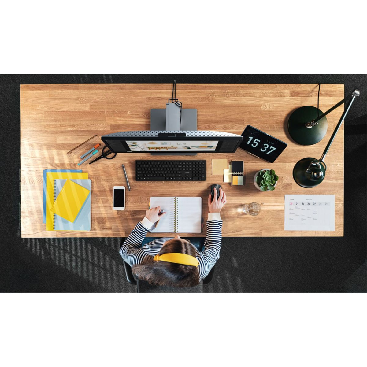 Draufsicht einer Person an einem Schreibtisch mit Computer, Telefon und Notizbüchern. Eine Lampe, eine Uhr und ein Kalender sind ebenfalls sichtbar.