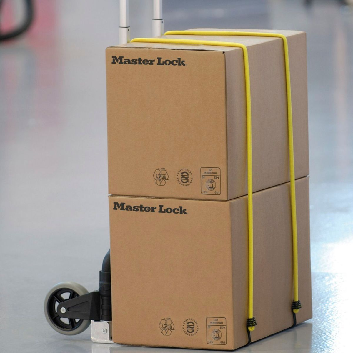 Due scatole di cartone impilate su un carrello portabagagli, fissate con corde elastiche gialle, etichettate 'Master Lock'.