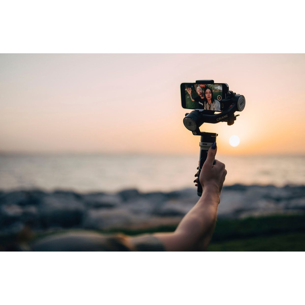Una persona sosteniendo un teléfono inteligente en un cardán, capturando una puesta de sol sobre el mar.