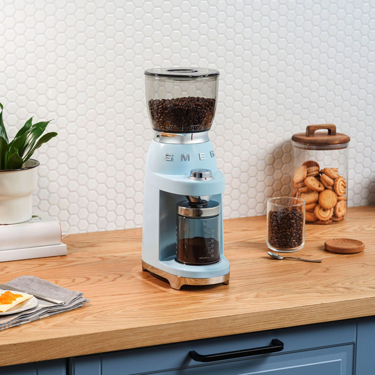 Un moulin à café bleu clair avec des grains de café à l'intérieur. Un pot de biscuits et un verre de grains sont à proximité.