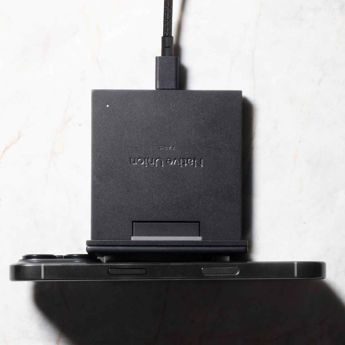 Ein schwarzes Native Union Ladegerät mit Telefon steht auf einer weißen Marmoroberfläche. Ein schwarzes Kabel ist angeschlossen.